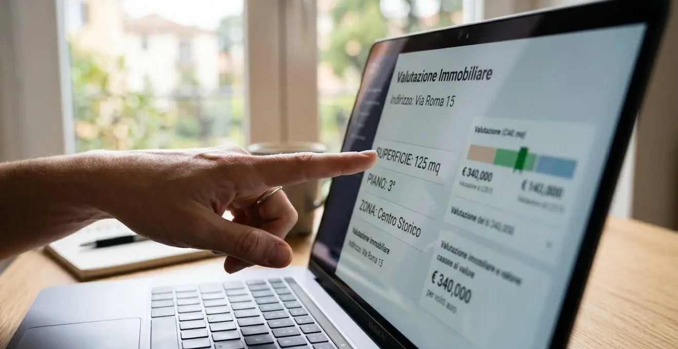 Estremo primo piano di uno schermo di laptop che mostra l'interfaccia di una piattaforma di valutazione immobiliare con campi in italiano, mano che indica un valore, superficie di scrivania moderna